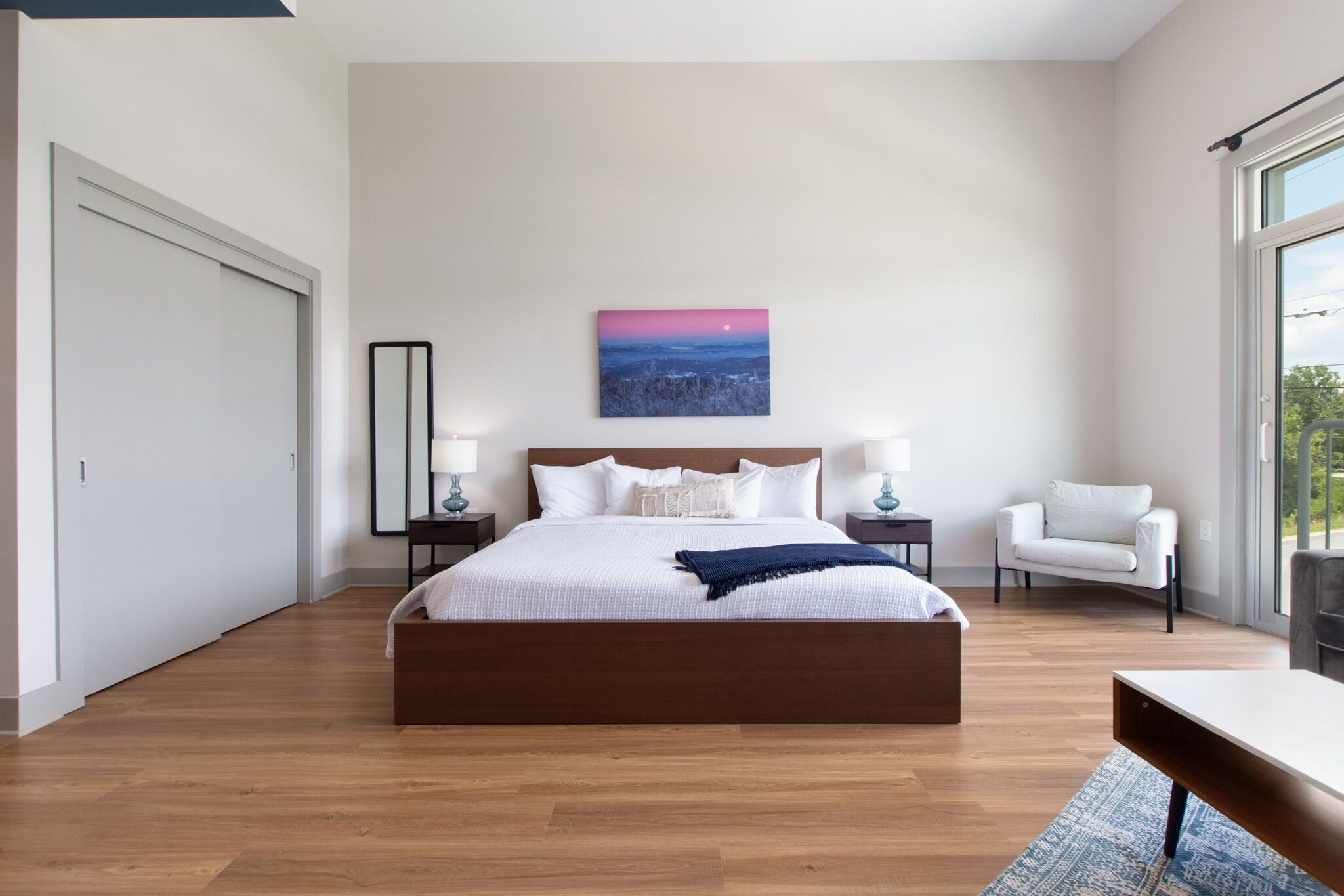 Une chambre à coucher moderne avec un grand lit, des tables de chevet, une chaise et un tableau au mur.
