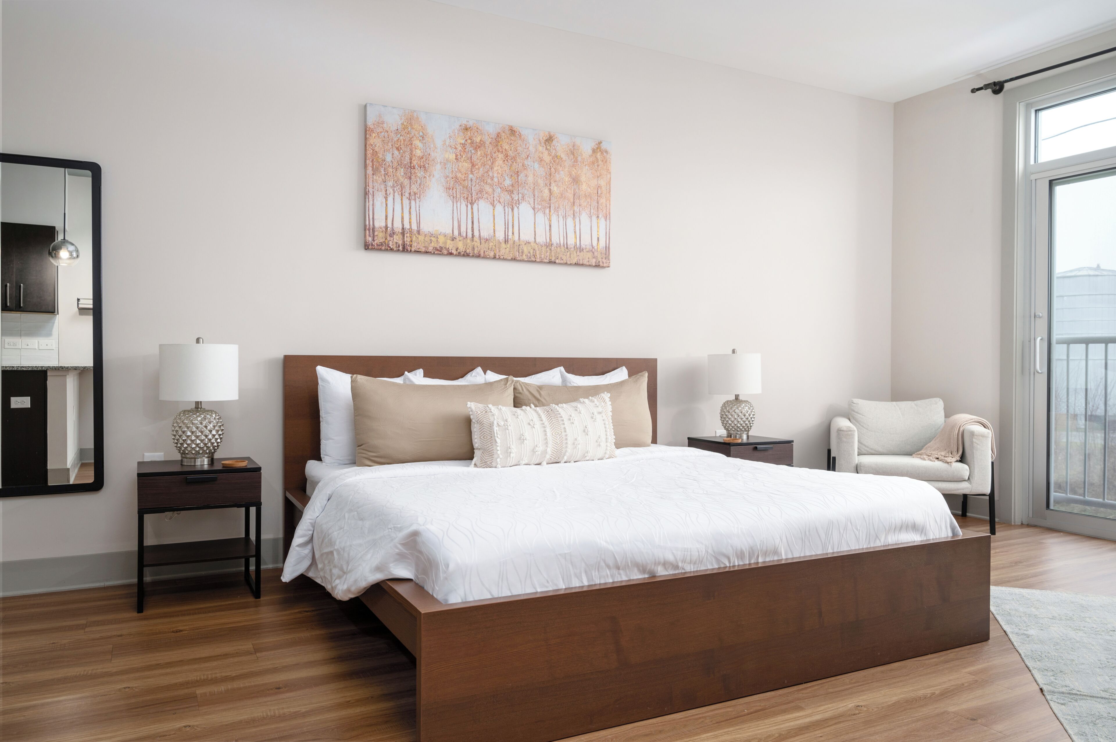 A modern bedroom with a large bed, two bedside tables, a chair, a painting on the wall, and a large window.