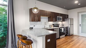 A modern kitchen with dark wooden cabinets, a granite countertop, stainless steel appliances, and a large window with a view of greenery. - River Row Flats (Asheville)