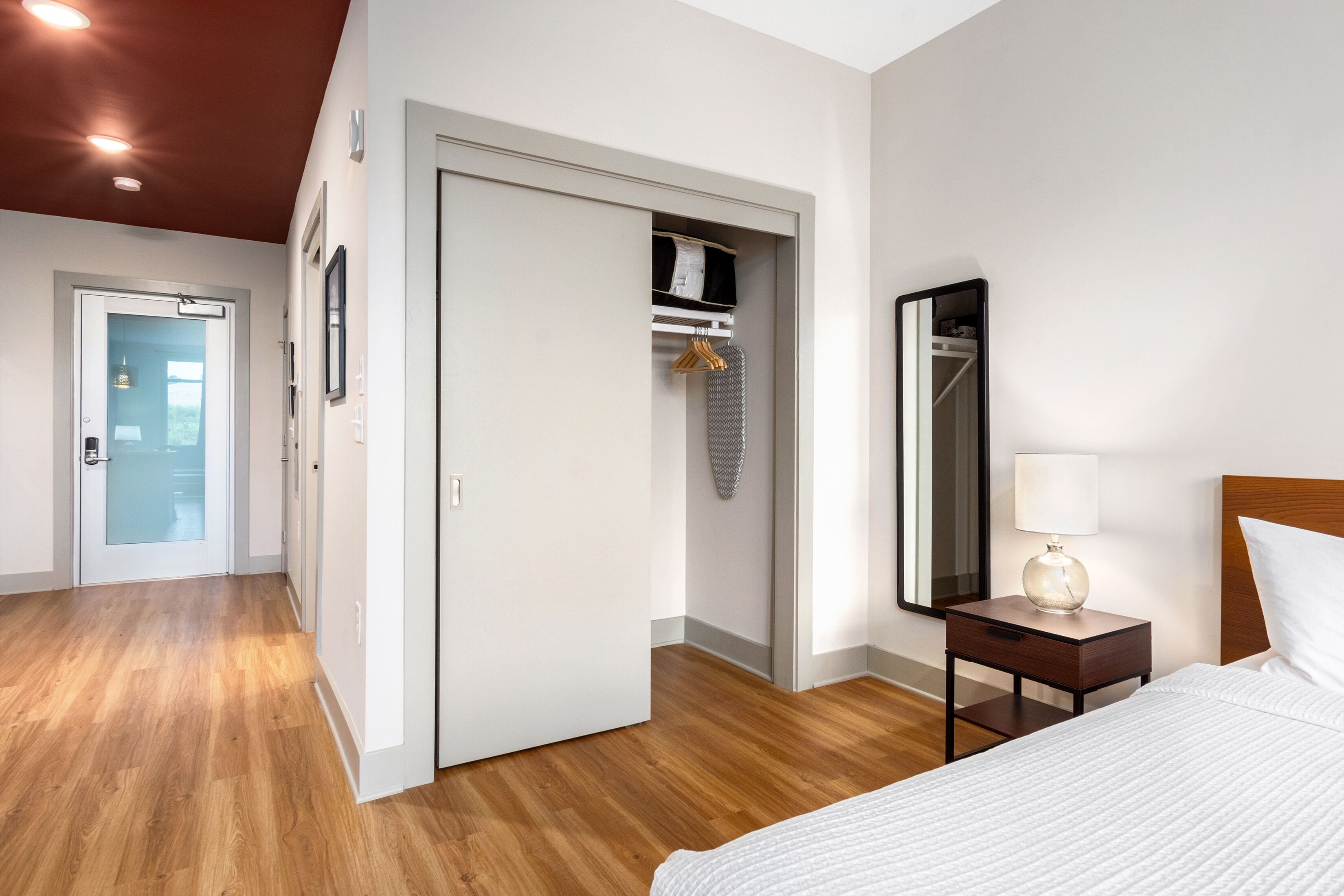 A hotel room with a wooden floor, a bed with white bedding, a nightstand with a lamp, a wardrobe, and a mirror.