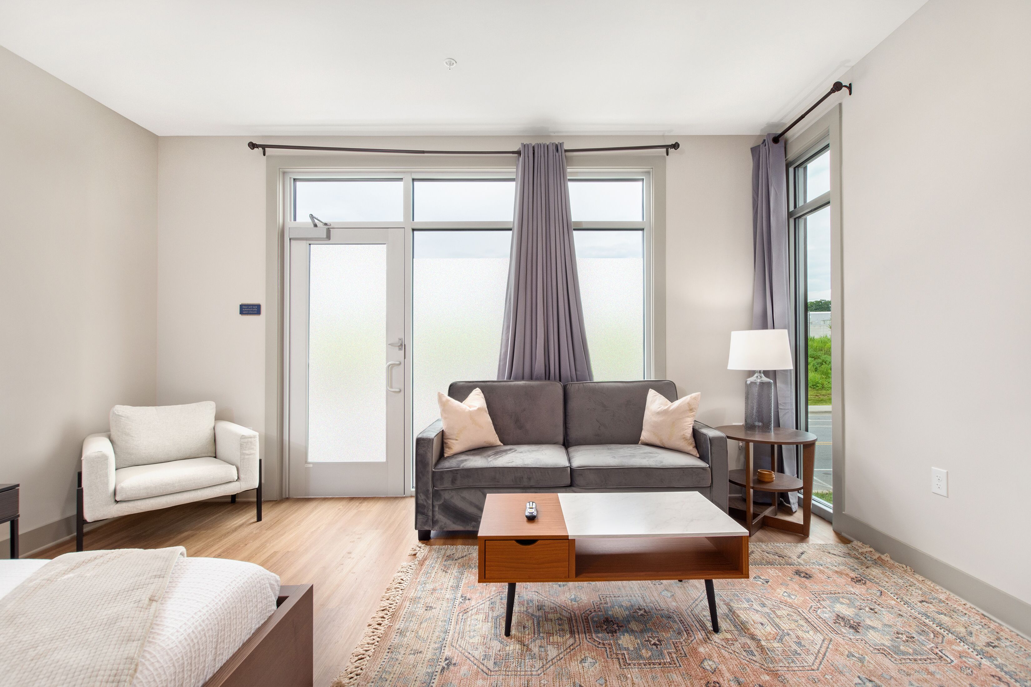 A modern hotel room with a sofa, a small table, a chair, and a large window with curtains.