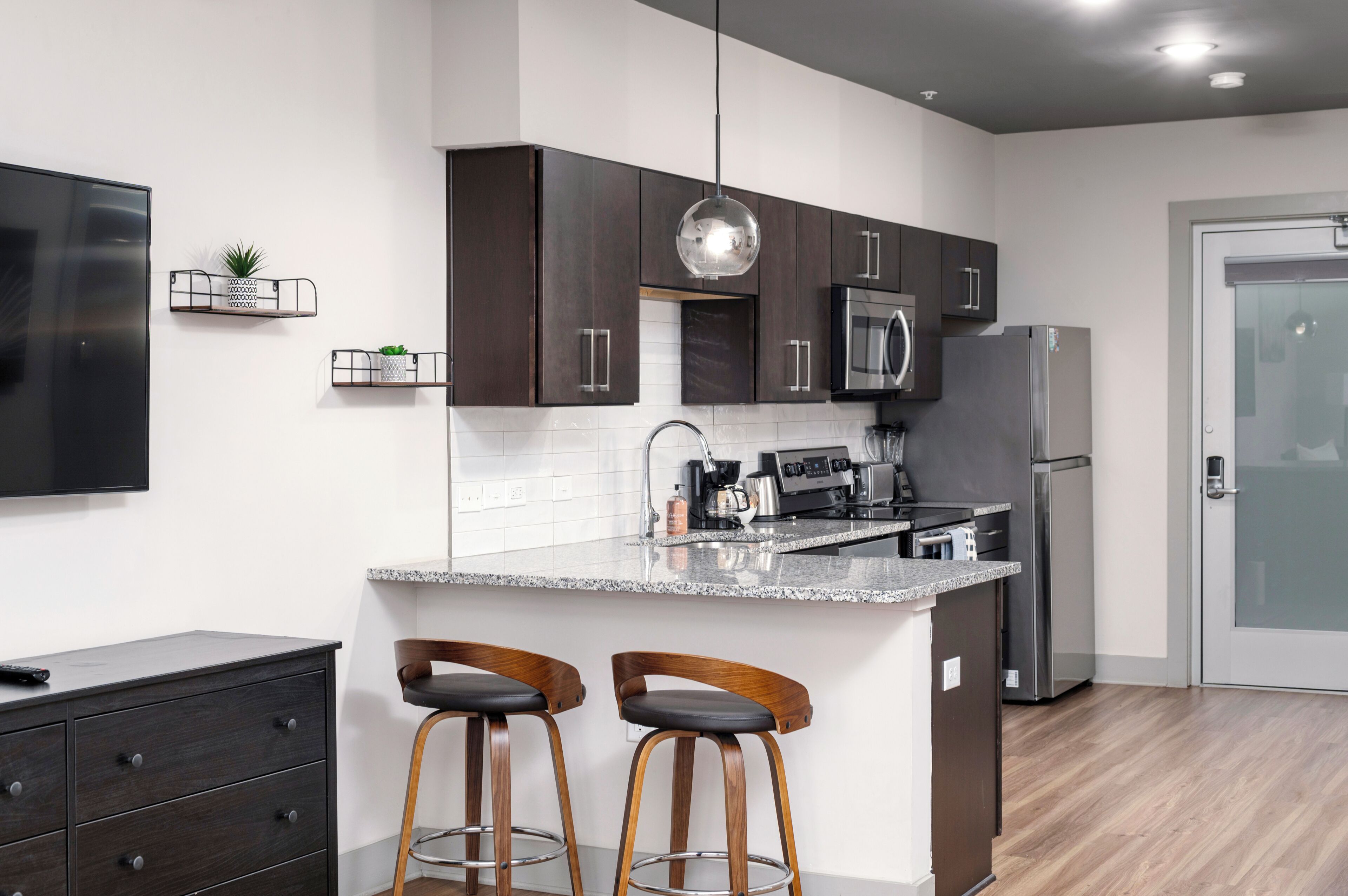 A modern kitchen with dark cabinets, a stainless steel refrigerator, a microwave, and a coffee machine. There are two wooden bar stools with metal legs.