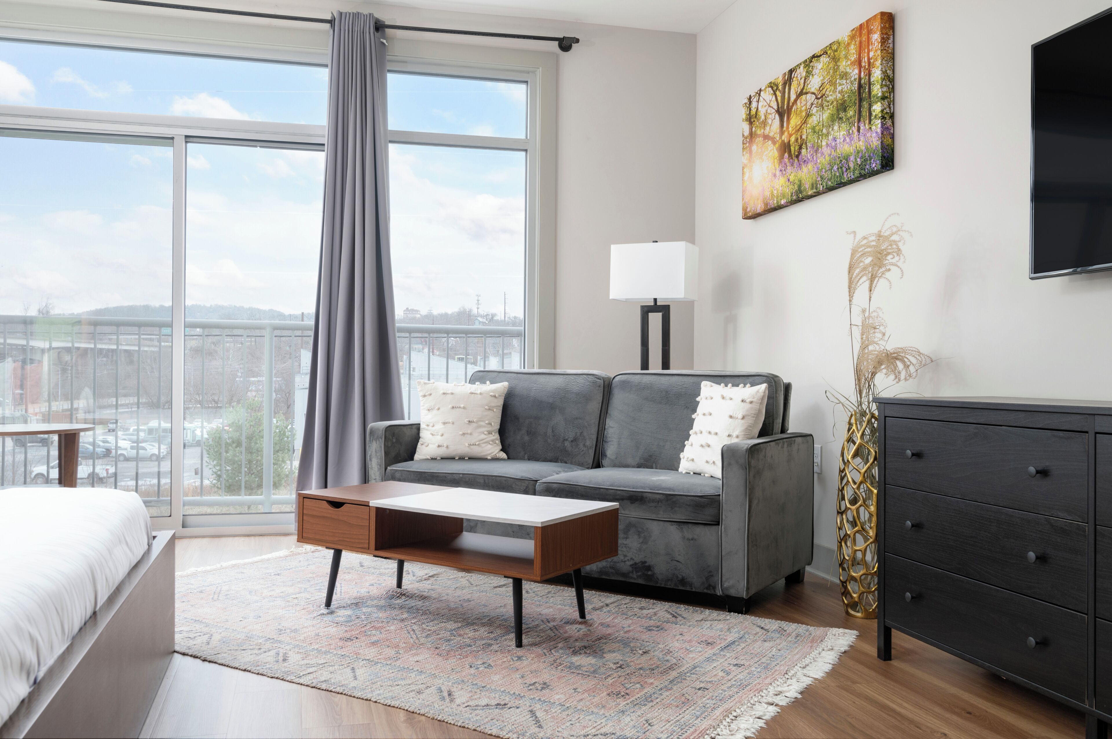 A modern living room with a grey sofa, a wooden coffee table, and a view of the city through large windows.