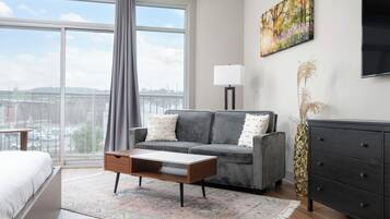 A modern living room with a grey sofa, a wooden coffee table, and a view of the city through large windows.