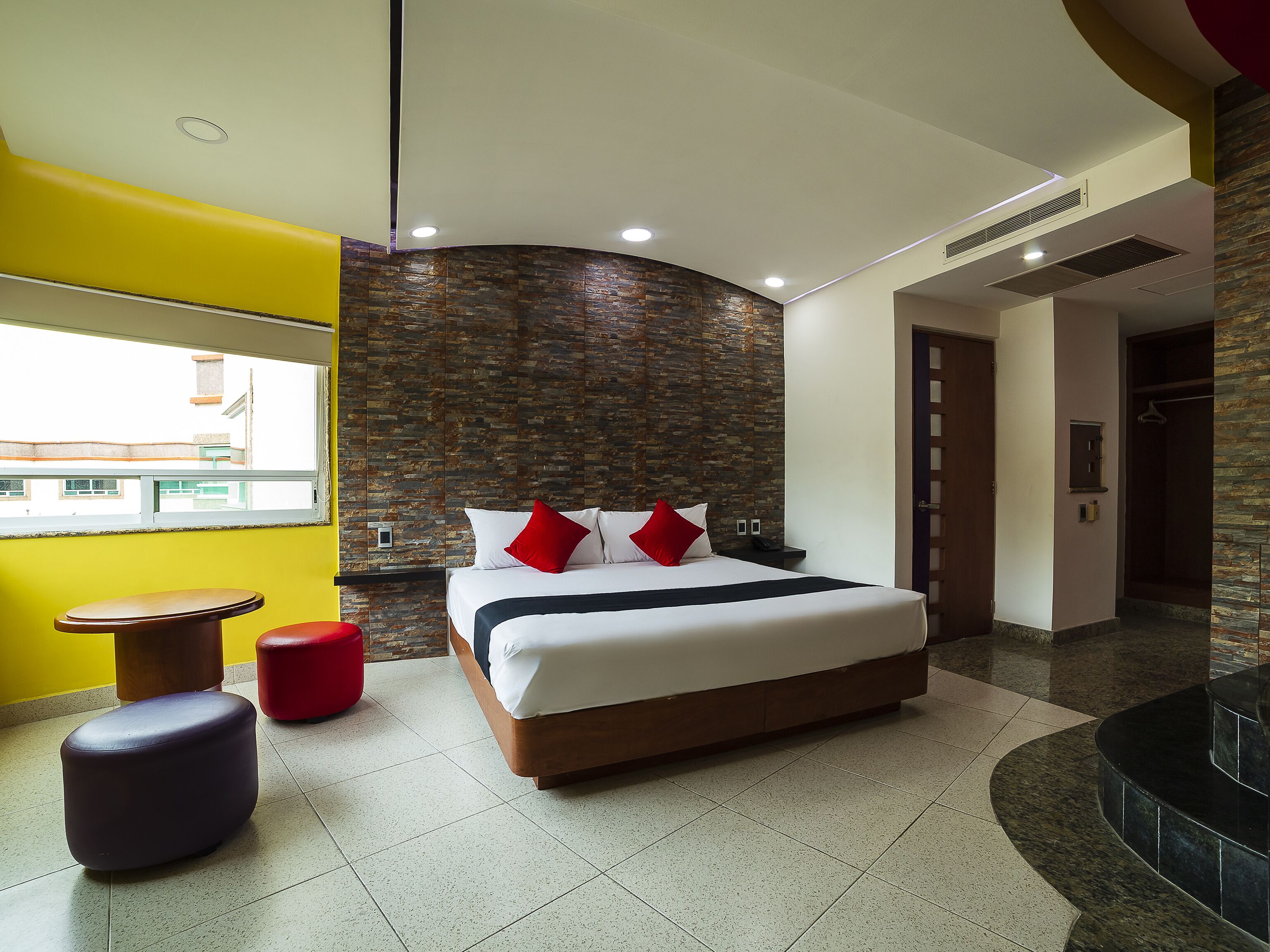 Camera d'albergo moderna con un letto grande, un tavolino, un poggiapiedi rosso e vista sulla città attraverso una finestra.
