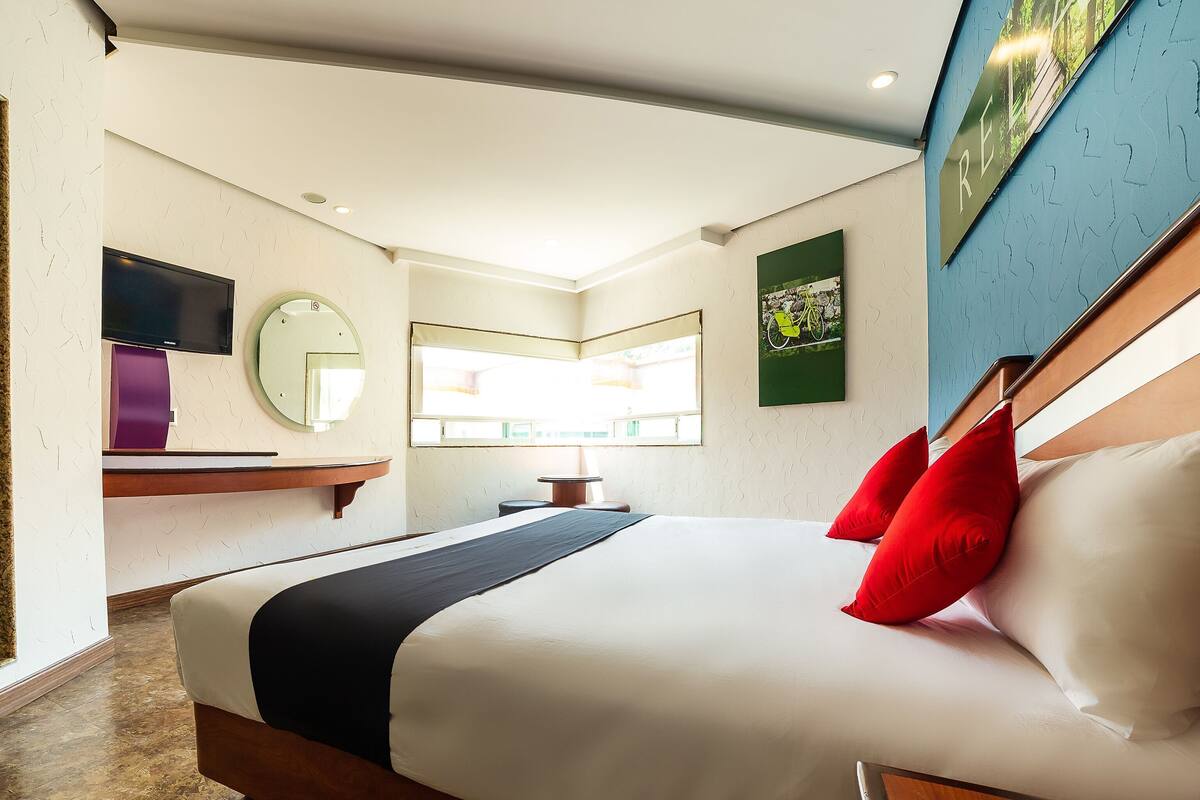 A hotel room with a bed, a bedside table, a mirror, a window, and a wall-mounted artwork.