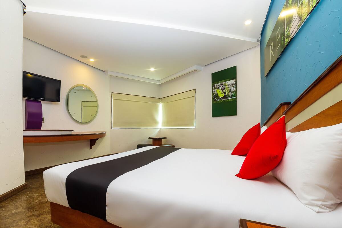 A hotel room with a bed, a desk, a television, a mirror, and a window with blinds.