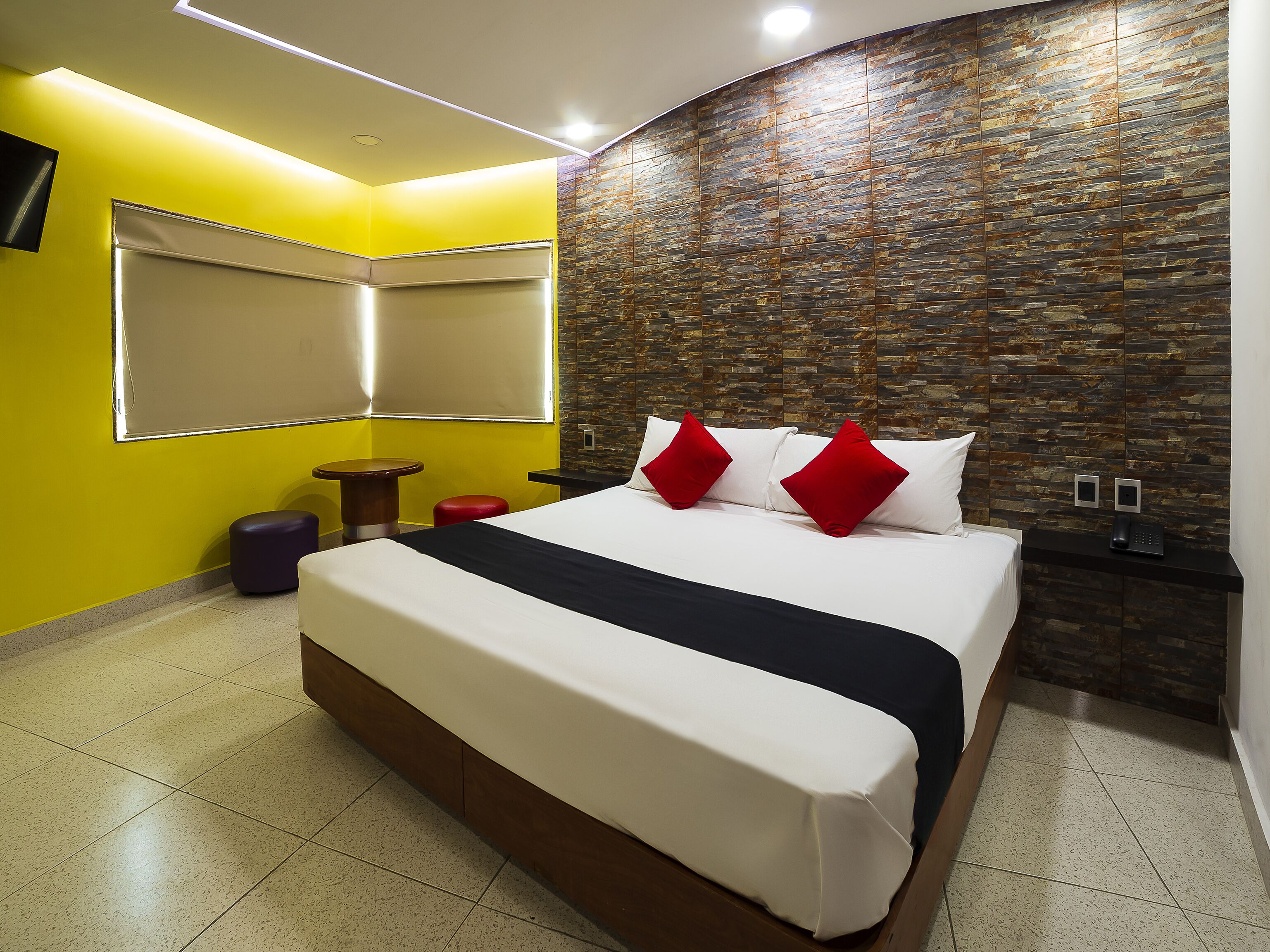 Una habitación de hotel moderna con una cama grande, una mesa redonda pequeña y un taburete rojo.