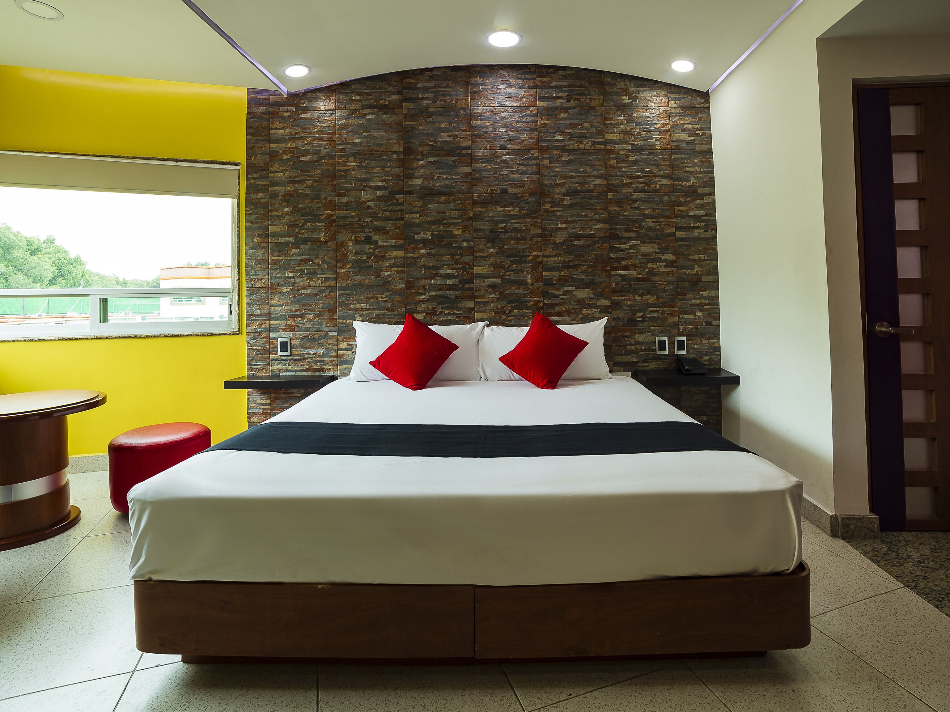 A hotel room with a large bed, two red pillows, a nightstand with a phone, and a window with a view of buildings.