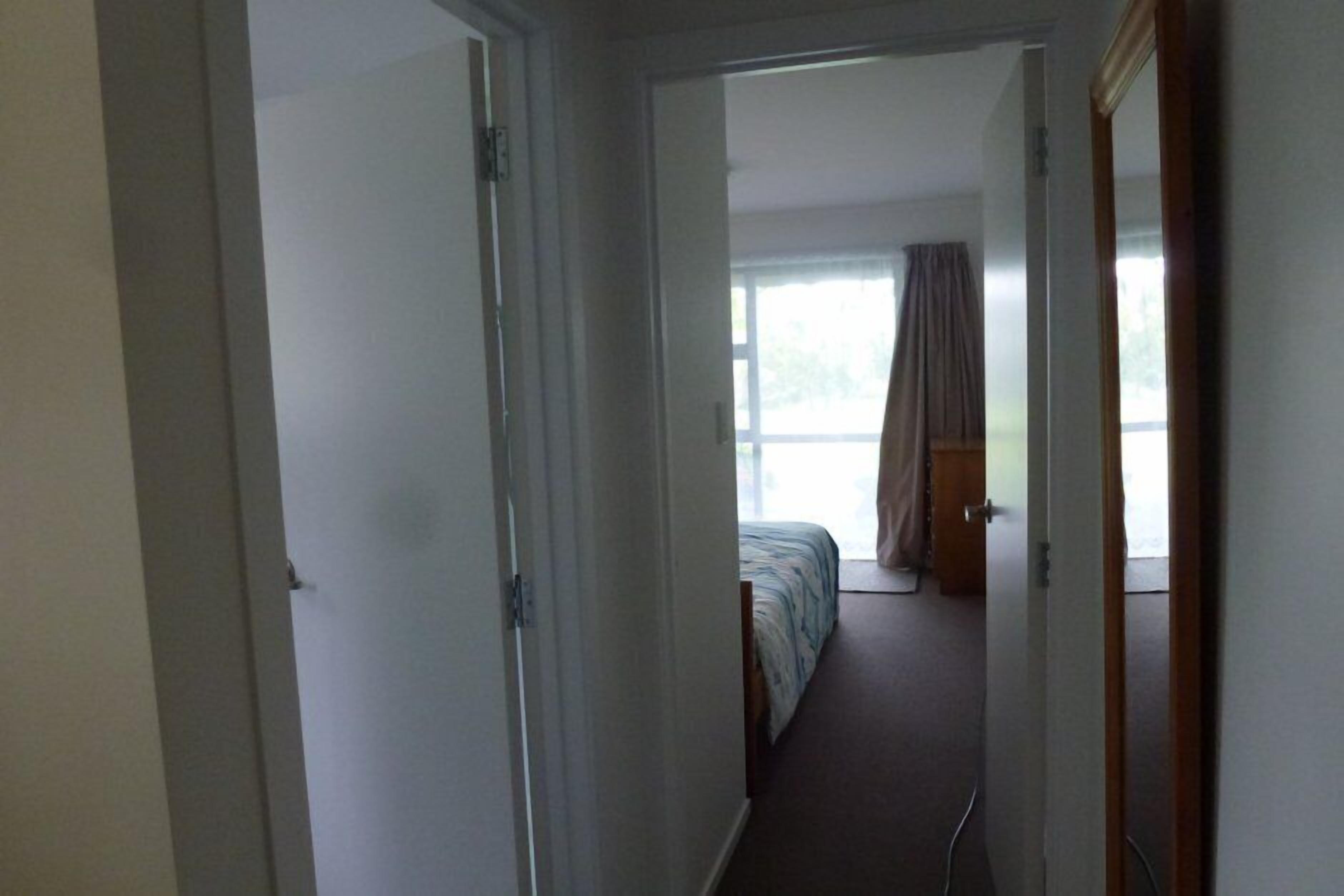 Een smalle gang die leidt naar een slaapkamer met een bed, een raam met gordijnen en een deur.