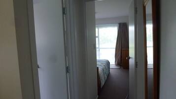 Een smalle gang die leidt naar een slaapkamer met een bed, een raam met gordijnen en een deur.