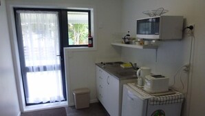 A small kitchen with a microwave, coffee maker, and a refrigerator. There is a window with curtains and a door leading outside. - Monty's self-contained unit (Kawhia)
