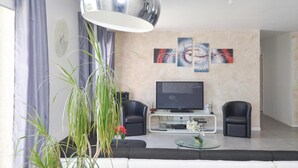 Uma sala de estar moderna com uma TV de tela plana, duas poltronas de couro preto, uma mesa de centro de vidro e um espelho grande acima da TV.
