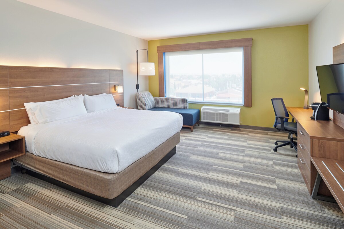 A hotel room with a large bed, a desk with a chair, a TV, a window, and a wall-mounted lamp.
