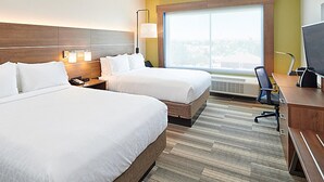 Quarto de hotel com duas camas, uma escrivaninha, uma cadeira, uma luminária e uma janela grande.