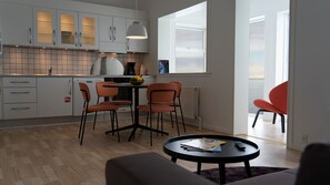 A modern kitchen with white cabinets, a dining area with two orange chairs, a round table, and a view of the sea through a large window.