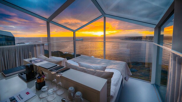 A modern room with a glass ceiling, a bed, a desk, and a view of the sunset over the water. - Hotel Arctic (Ilulissat)