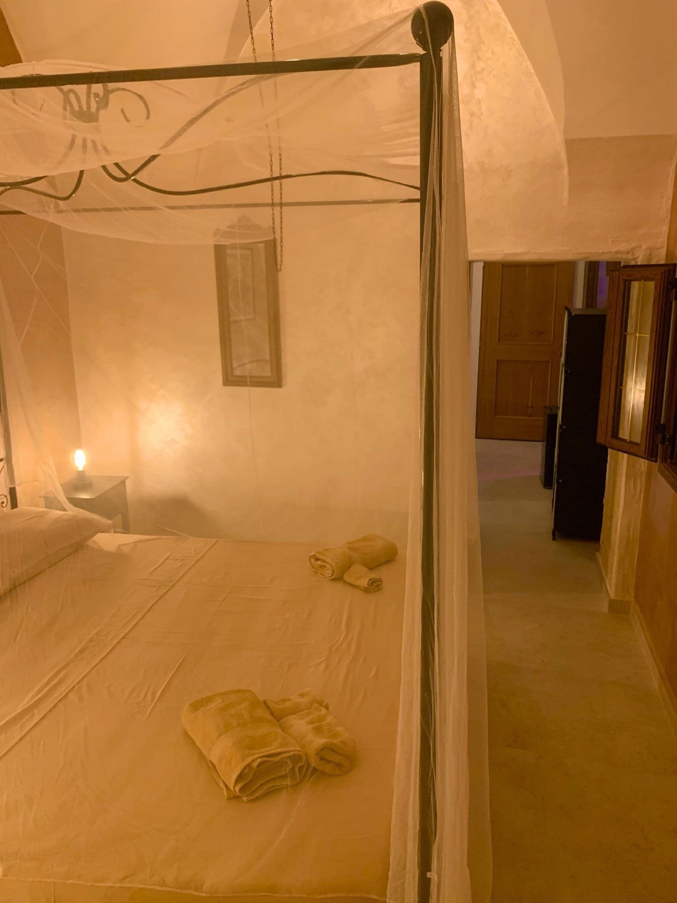 A bedroom with a four-poster bed, a bedside table with a lamp, and a rolled towel on the bed.