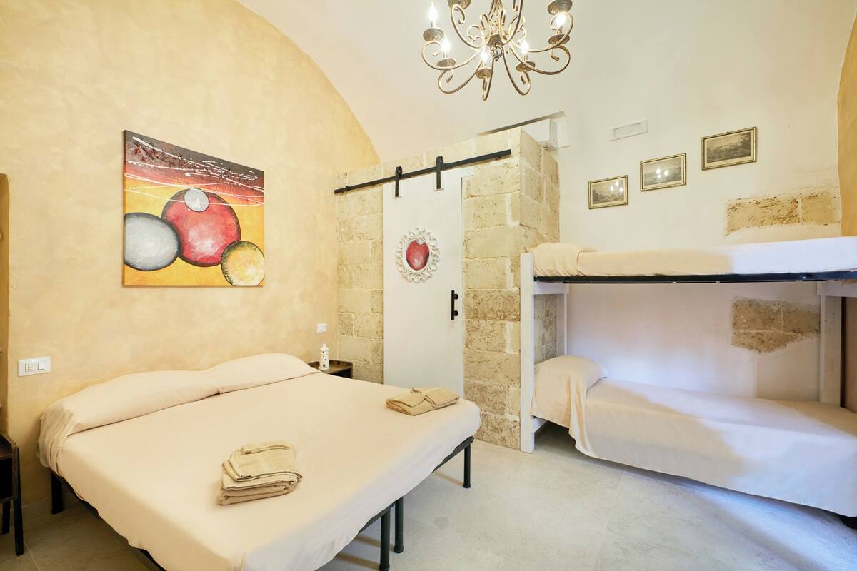 A hotel room with a bunk bed, a single bed, a painting on the wall, a chandelier, and a small nightstand.