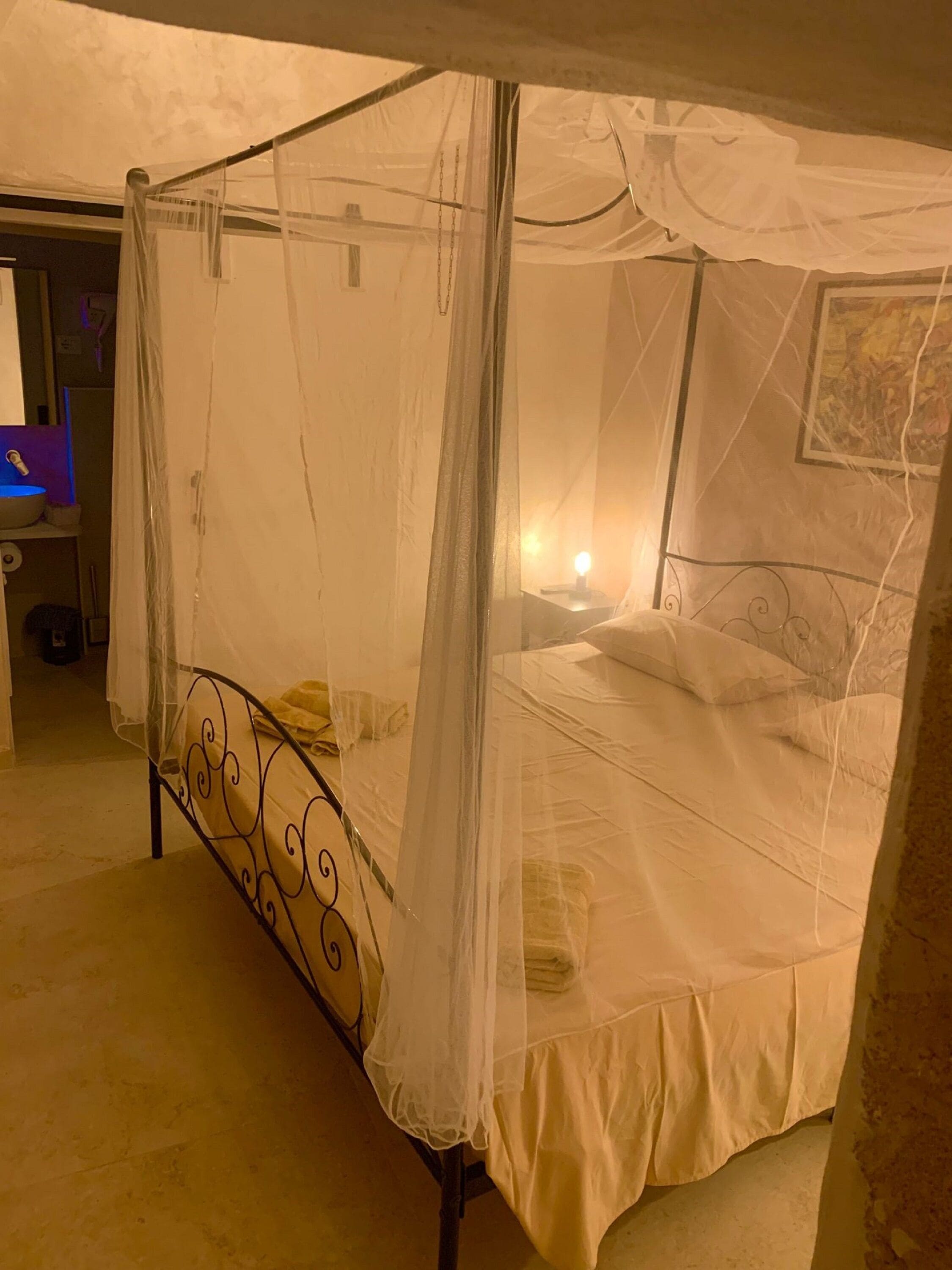 A four-poster canopy bed with a white bedspread and pillows, a bedside table with a lamp, and a wall-mounted picture.