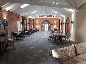 A spacious indoor dining area with brick walls, multiple tables and chairs, and a high ceiling with skylights.