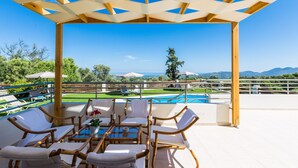 A covered outdoor seating area with a pool view, wooden pergola, and white lounge chairs. - Anatoli Villa, a Retreat with Private Pool & BBQ (Rethymno)