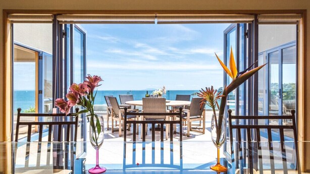 Una terraza abierta con mesa de vidrio, sillas y jarrones con flores, con vistas a un área de comedor y al mar.