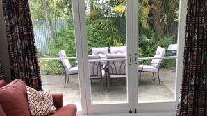 A living room with a red sofa, patterned cushions, a large patterned rug, and a sliding glass door leading to an outdoor patio with a table and chairs. - Walk to the wharf and village (Mapua (Ruby Bay))