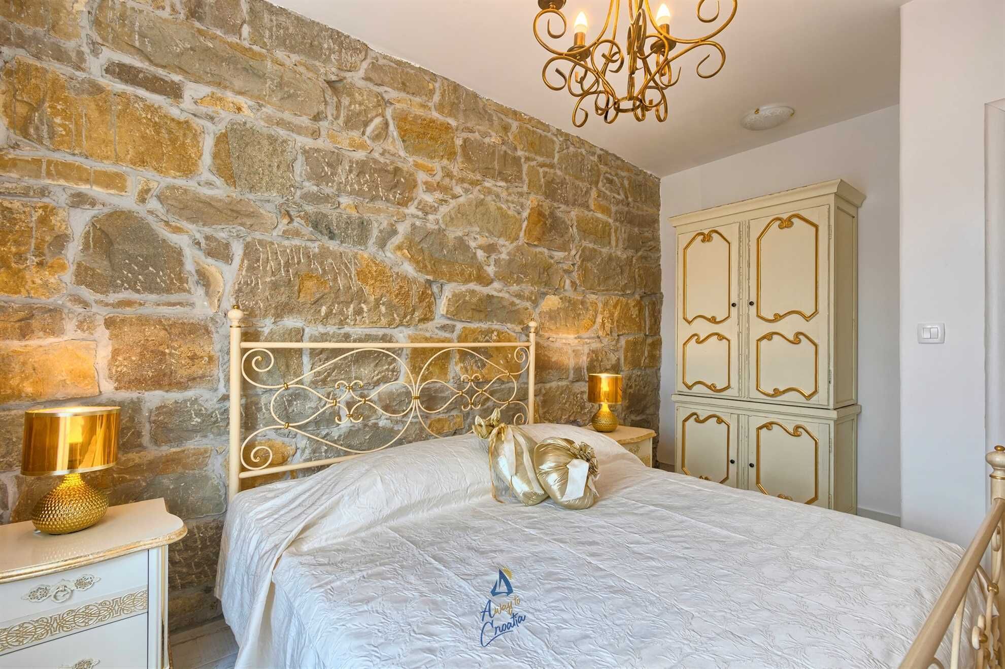 Un dormitorio con pared de piedra, una cama con cabecera decorativa, una mesita de noche con lámpara y un armario con espejo.