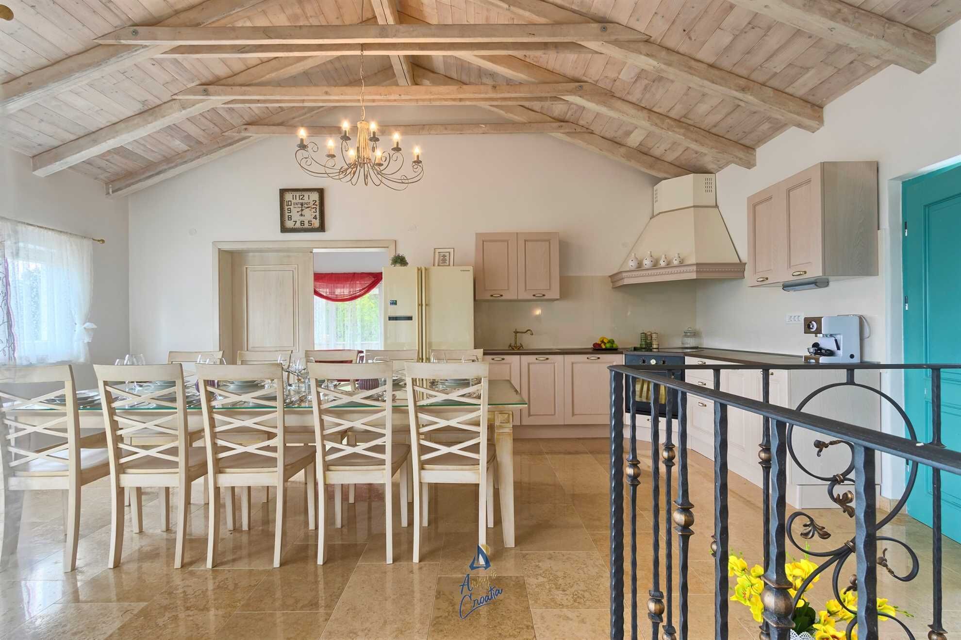 Une salle à manger spacieuse agrémentée d’un lustre, une cuisine aux meubles de couleur claire et un balcon doté d’une balustrade.