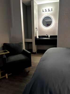 A hotel room with a bed, a black armchair, a round mirror with lights, a black vanity with a sink, and a white towel on the wall.