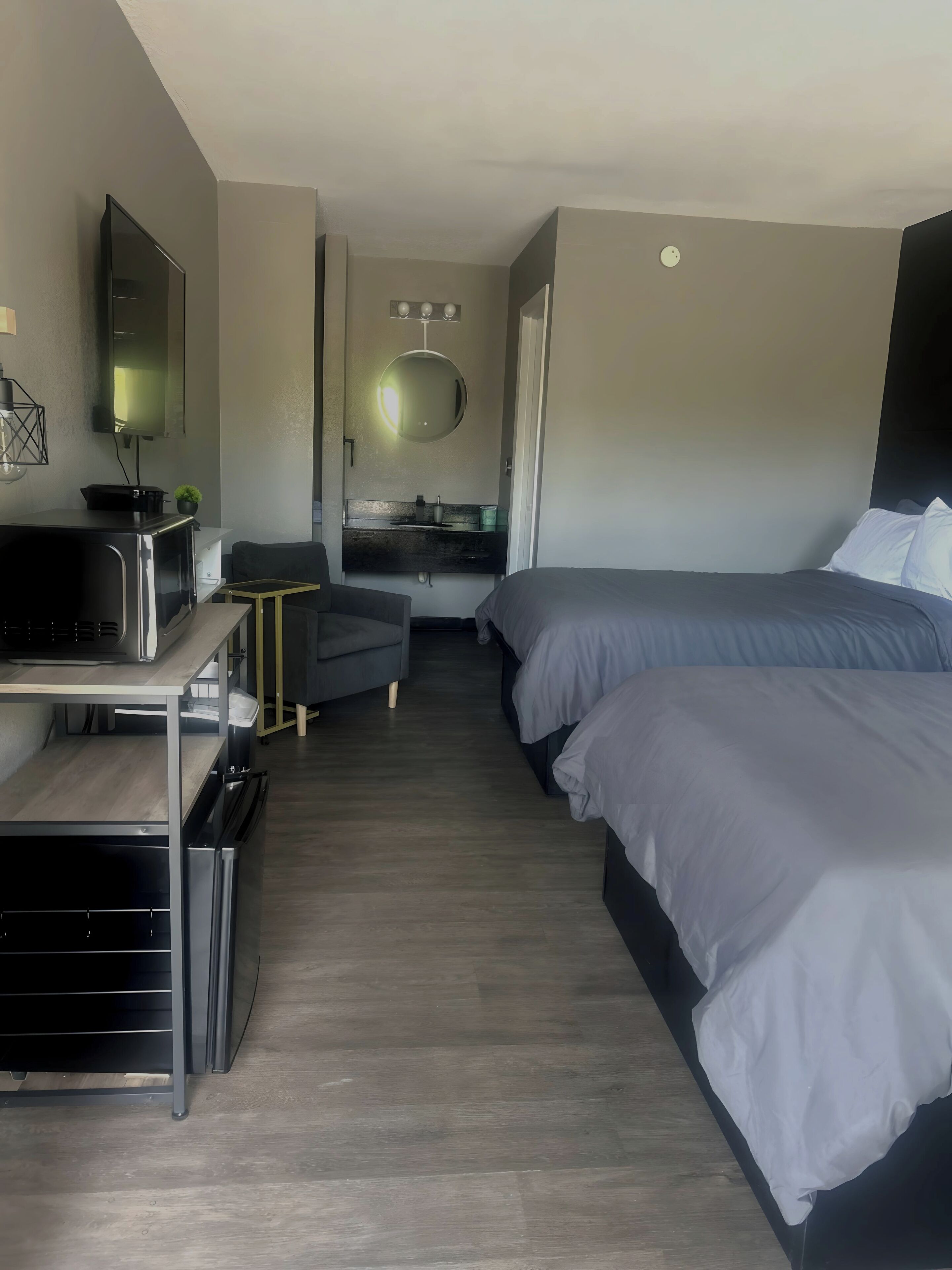 A modern hotel room with a desk, two beds, a small kitchenette, and a mirror.