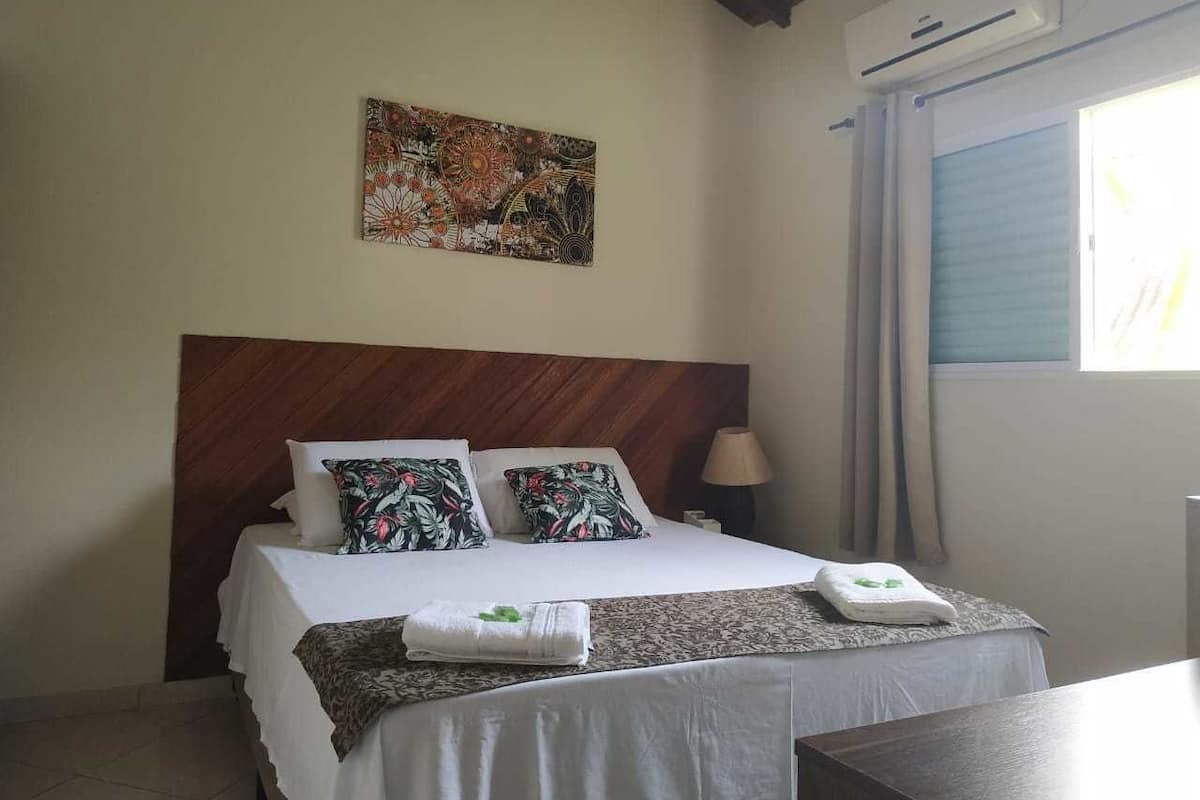 Une chambre d’hôtel avec un lit, une tête de lit, un climatiseur fixé au mur, une fenêtre avec des rideaux et un tableau accroché au mur.
