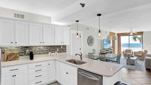 Eine moderne Küche mit weißen Schränken, einem Edelstahl-Geschirrspüler und einer großen Kochinsel. Ein Essbereich mit Tisch und Stühlen lädt zum Verweilen ein, und ein Wohnzimmer bietet einen Blick auf den Ozean.