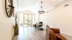 Uma sala de estar ampla com piso de madeira, um sofá de couro marrom, uma mesa de jantar com cadeiras e um candelabro.