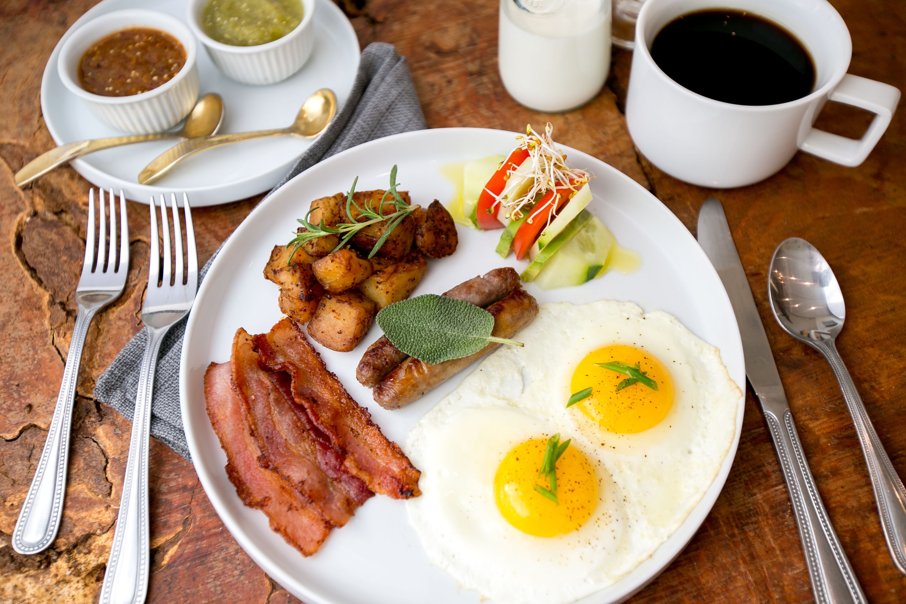 Prato de pequeno-almoço com bacon, ovos, salsichas e legumes sobre uma mesa de madeira.