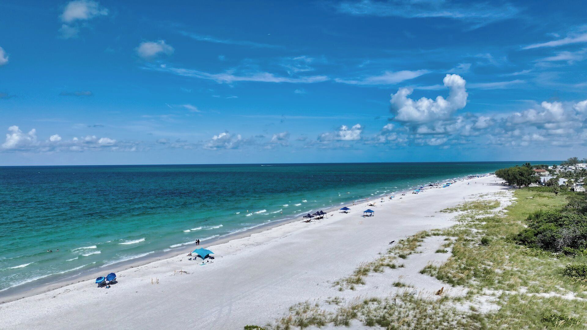 A beach with white sand, clear blue water, and a few umbrellas.
