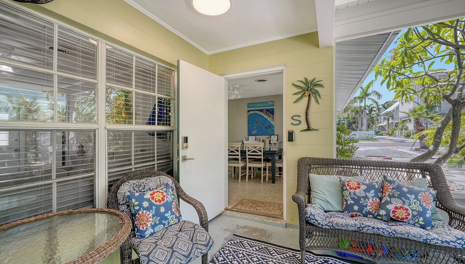 A room with wicker furniture, a door leading to another room, and a view of a street with palm trees.