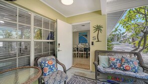 Una habitación con muebles de mimbre, una puerta que da a otra habitación y vista a una calle con palmeras.