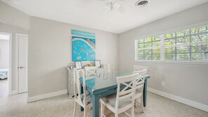 Un comedor con mesa de vidrio y sillas blancas, una obra de arte azul en la pared, y una ventana con persianas.