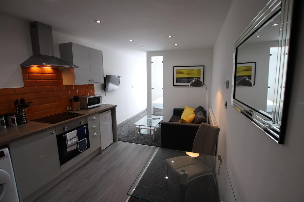 A modern kitchen with a microwave, oven, and sink. There is a glass table and a sofa with a yellow pillow. A large mirror is on the wall.