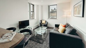 A modern living room with a glass coffee table, a dark grey sofa, a wooden dining table, and a flat-screen TV. - Halifax House Apartments (Halifax)
