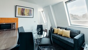A modern living room with a dining table, chairs, a gray sofa with yellow cushions, a microwave, and a large window with a view of buildings. - Halifax House Apartments (Halifax)