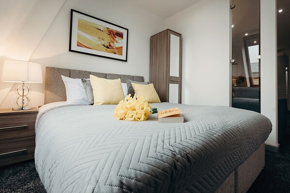 A neatly made bed with a quilted grey bedspread, a bouquet of yellow flowers, and a book on the bed. - Halifax House Apartments (Halifax)
