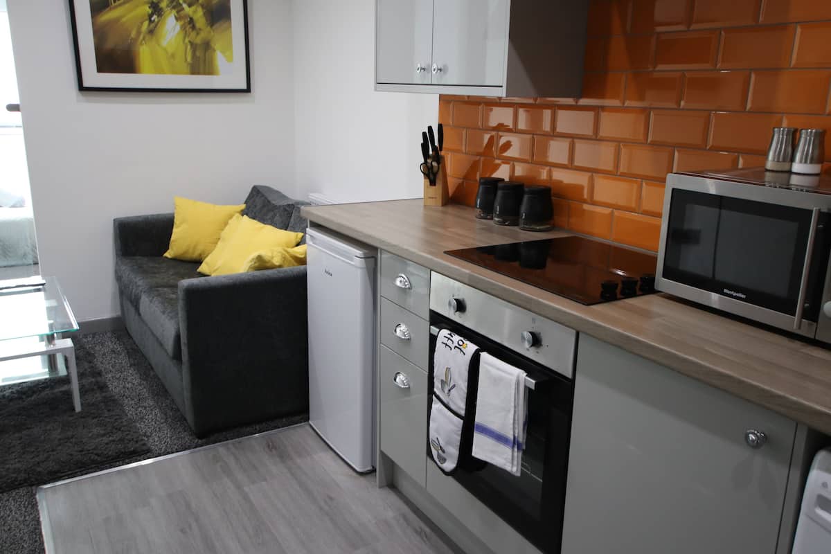 A compact kitchen with a microwave, oven, and a small refrigerator. There is a grey sofa with yellow cushions in the adjacent living area.