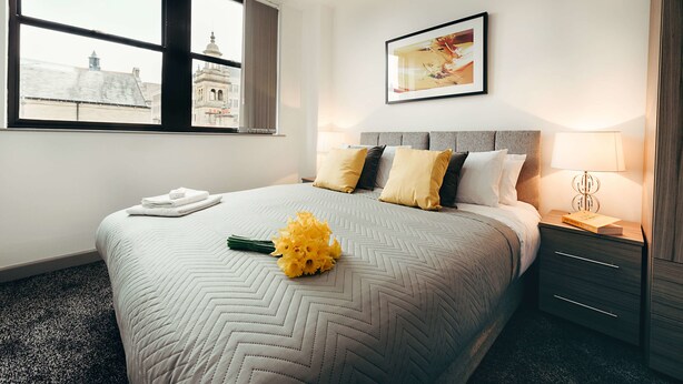 A hotel room with a large bed, a nightstand, a lamp, and a window with a city view. - Halifax House Apartments (Halifax)