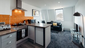 A compact kitchen with a built-in oven, a microwave, and a gas stove. There is a small refrigerator, a sink, and a wooden countertop. The kitchen is adjacent to a living area with a sofa and a television. - Halifax House Apartments (Halifax)