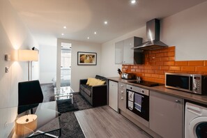 A modern kitchen with a microwave, oven, and washing machine. There is a glass table with a lamp, a sofa with cushions, and a picture on the wall. - Halifax House Apartments (Halifax)