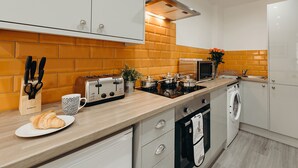 A modern kitchen with white cabinets, a brick backsplash, a toaster, a coffee maker, and a washing machine. - Halifax House Apartments (Halifax)