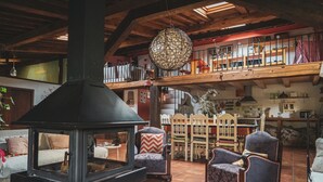 A rustic interior with a wood-burning stove, a dining area with chairs and a table, and a loft space with a balcony.
